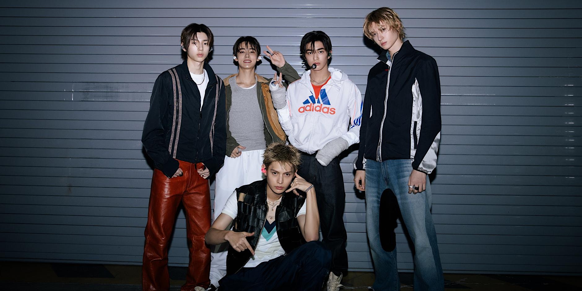 Five young men in streetwear pose in front of a gray metal shutter, with one seated in front wearing a black vest and white shirt.