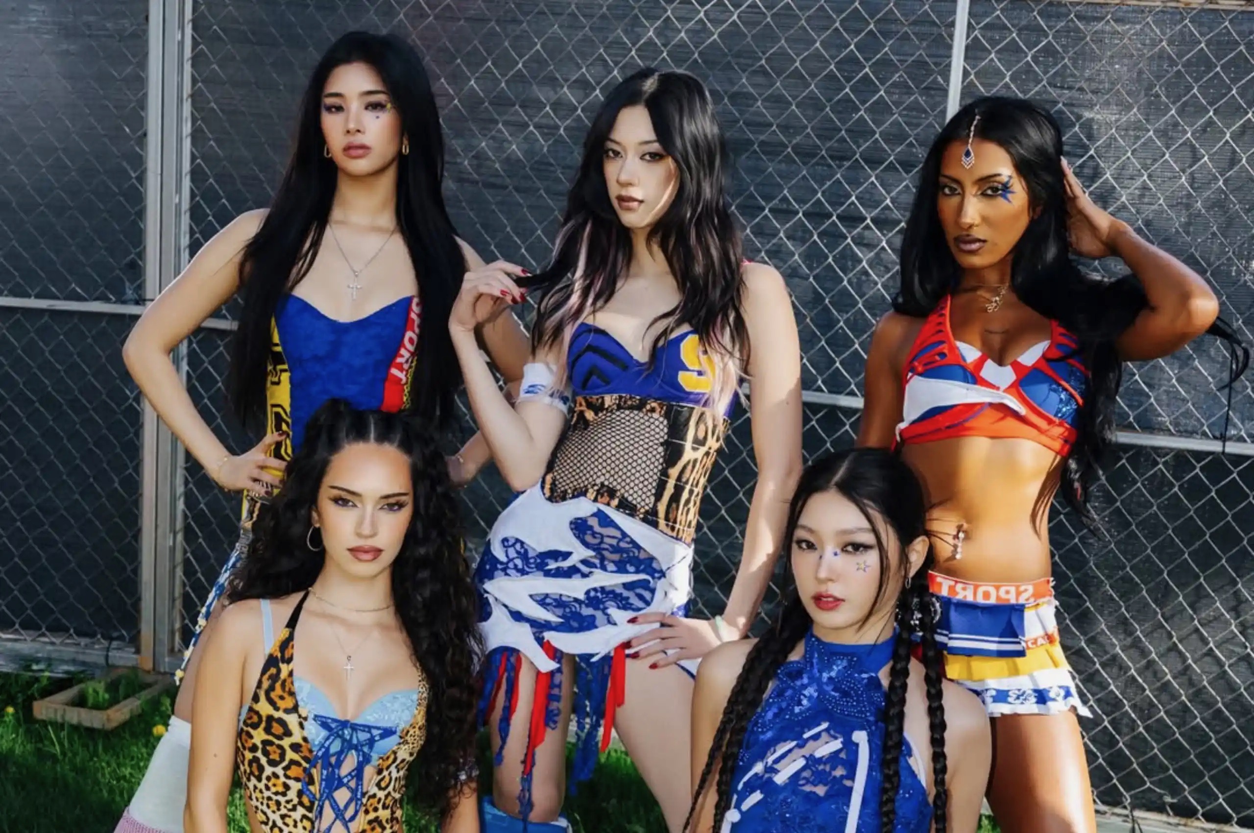 Group of women in colorful outfits posing together in front of a chain-link fence outdoors.
